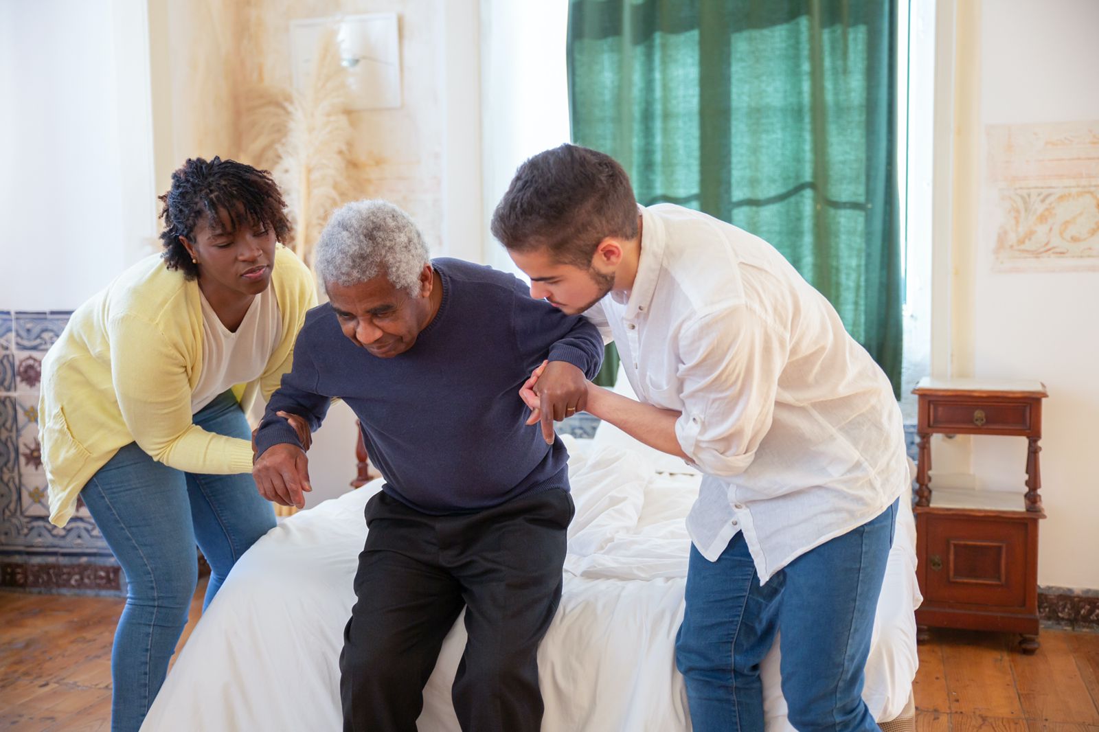 A male and female caregiver helping support an elderly man out of bed